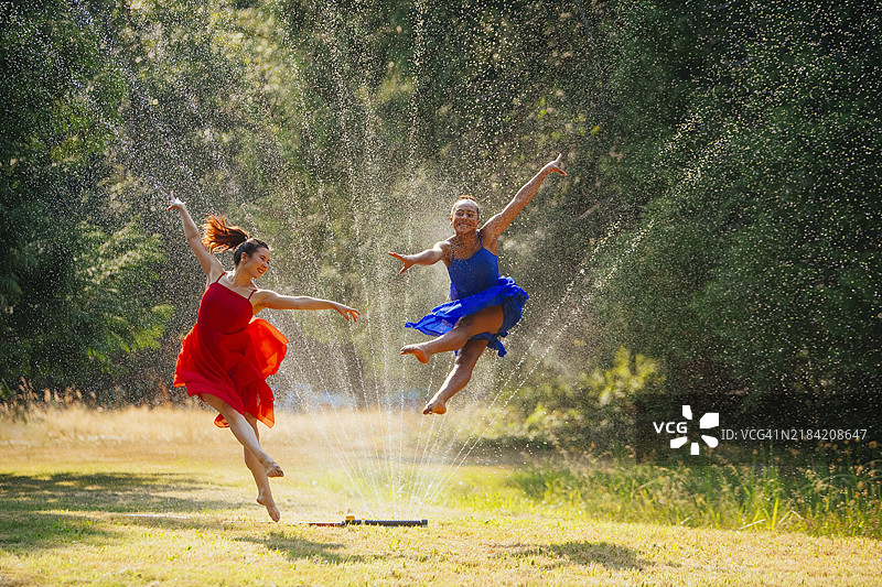 两个快乐的女人在阳光明媚的日子里，在草地上跳舞并穿越喷洒的水花。图片素材