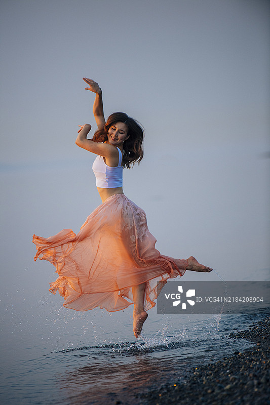在黄昏时分，穿着飘逸桃色裙子的快乐女性在海滩上跳舞图片素材