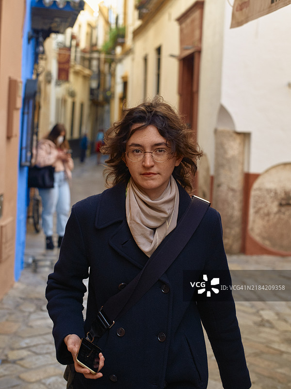 戴眼镜的休闲年轻人穿着海军蓝外套走在风景如画的狭窄街道上图片素材