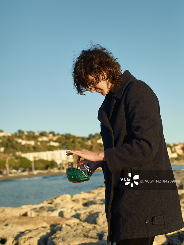 一个卷发的人穿着深色外套，站在海边，从一个绿色瓶子里倒出液体。图片素材