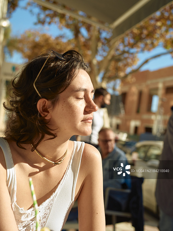 年轻女性在户外咖啡馆享受阳光，背景中有其他人坐着。图片素材