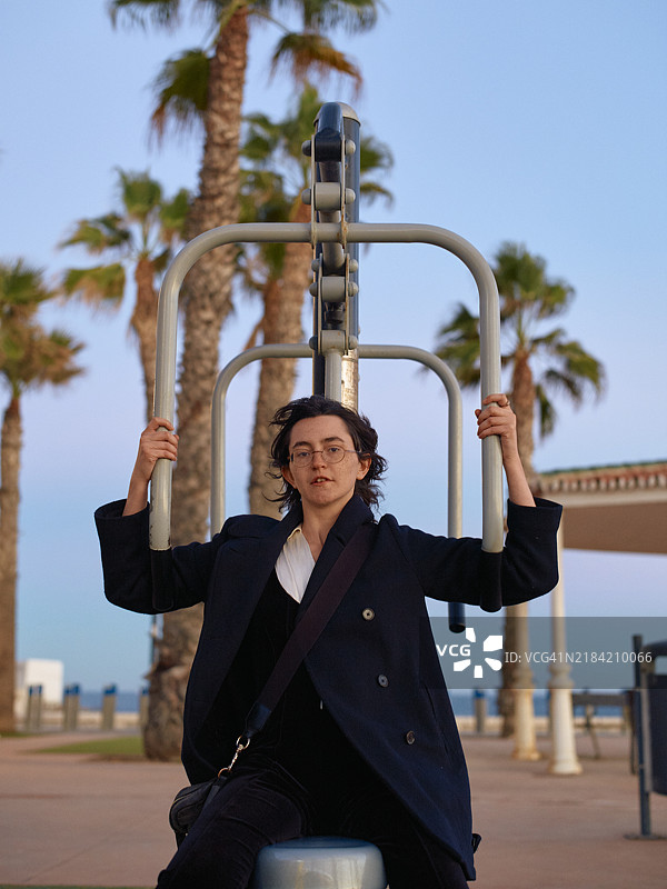一位戴眼镜的成年人在户外健身站进行过头推举，背景是棕榈树和黄昏时分的晴朗天空。图片素材
