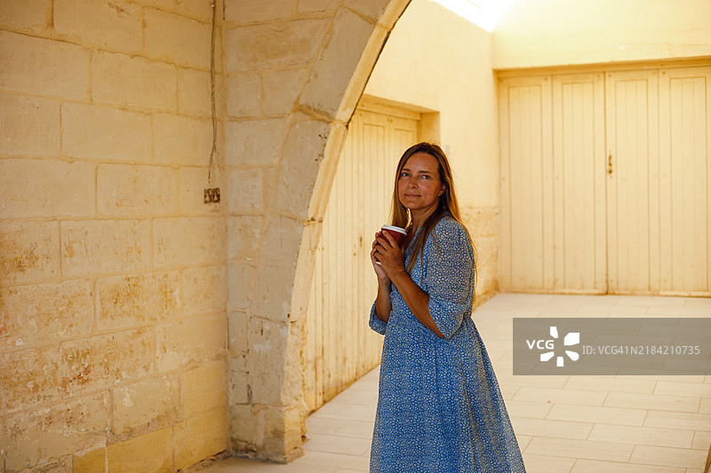 一位穿着蓝色花卉连衣裙的女性站在阳光明媚的地中海庭院中，她放松的姿势和温暖的微笑营造出宁静的氛围。非常适合旅行、生活方式和度假主题的图片。图片素材