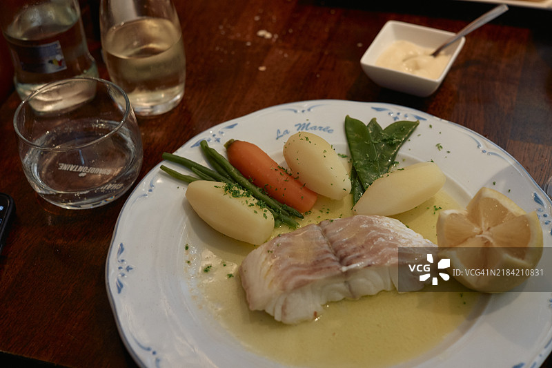一盘白盘上的水煮鱼配柠檬、煮土豆、胡萝卜和菠菜，旁边有一杯水和一杯白葡萄酒，摆放在木桌上。图片素材