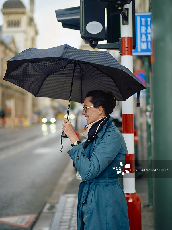 一位微笑的女性穿着蓝色外套，手持打开的雨伞，站在行人过街道上。图片素材