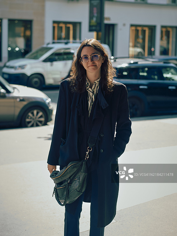 身穿海军蓝外套和条纹围巾的时尚人士，站在人行道上，肩背绿色手提包，佩戴太阳镜。图片素材