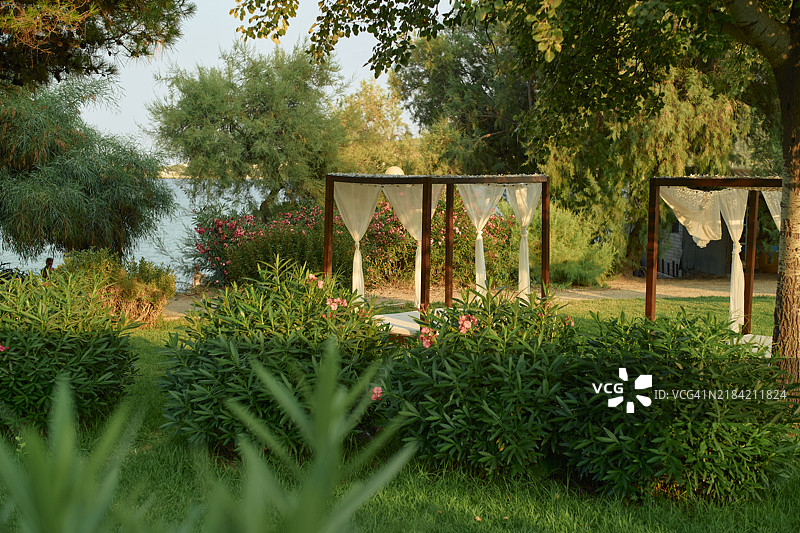 日出光线洒在宁静的花园里，背景是一个凉亭和湖泊，被丰盛的绿色植物和盛开的花卉环绕。图片素材