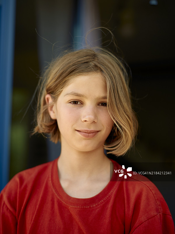 微笑的年轻女孩，短金发，穿着红色衬衫，站在模糊的背景前。图片素材