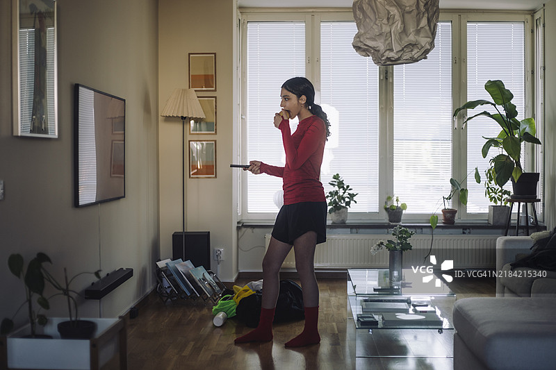 女孩侧身站在家里的客厅里，一边吃苹果一边看电视。图片素材