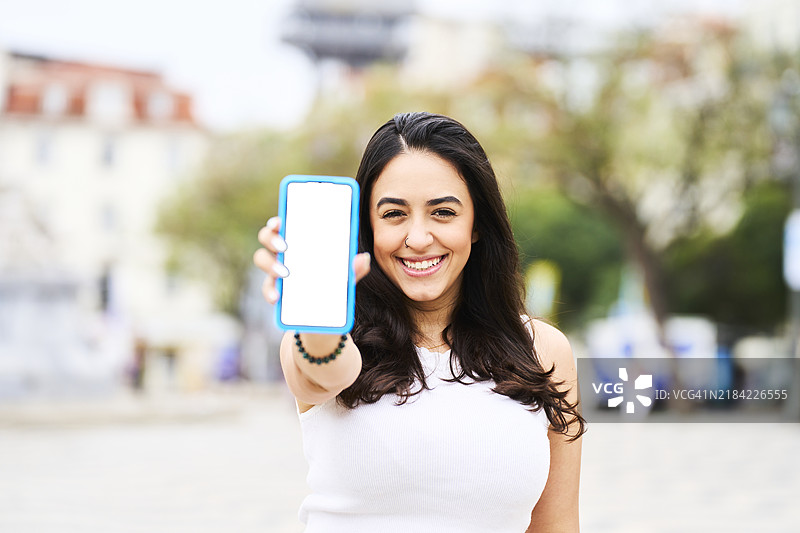 美丽的游客女性穿着T恤，微笑着展示手机屏幕，面对镜头，站在佩德罗四世广场，背景是圣胡斯塔电梯，正面视角。图片素材