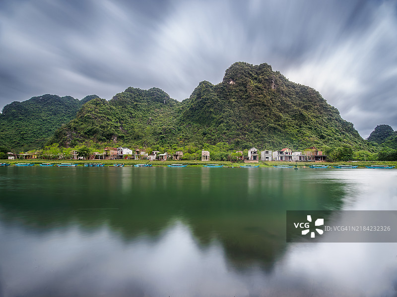 长时间曝光的喀斯特山脉景观，山上覆盖着森林和传统房屋，映照在河面上，河岸上停靠着小型木制蓝色旅游船，风牙村，风牙-凯邦国家公园 - 广平省 - 越南北部。图片素材