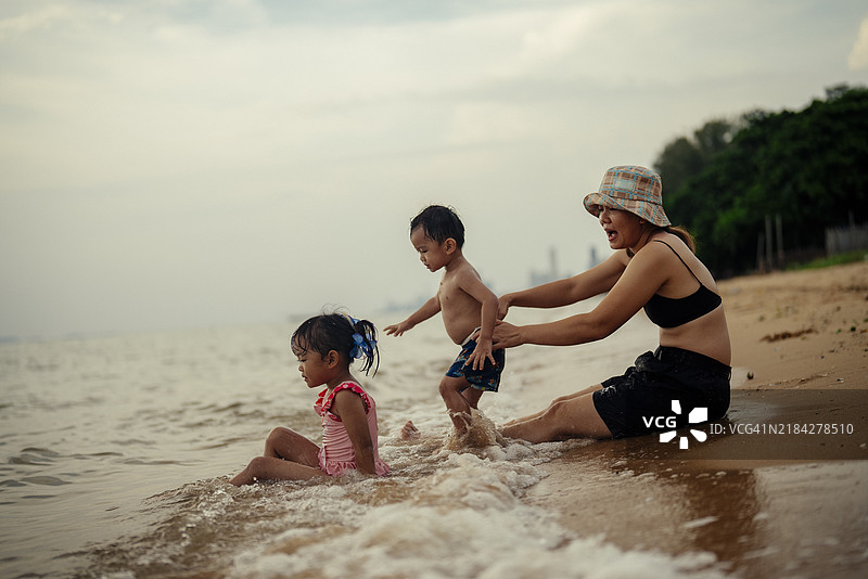 积极的情感：一位亚洲母亲和她的年轻孩子们在浅滩水中一起玩耍，戴着太阳帽，享受假期。图片素材