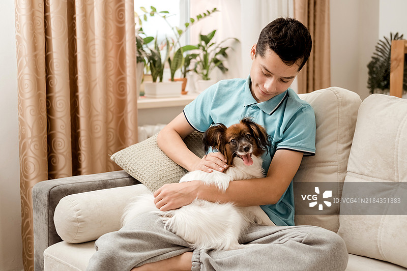 一个快乐的白人少年坐在沙发上，抱着他的狗。图片素材
