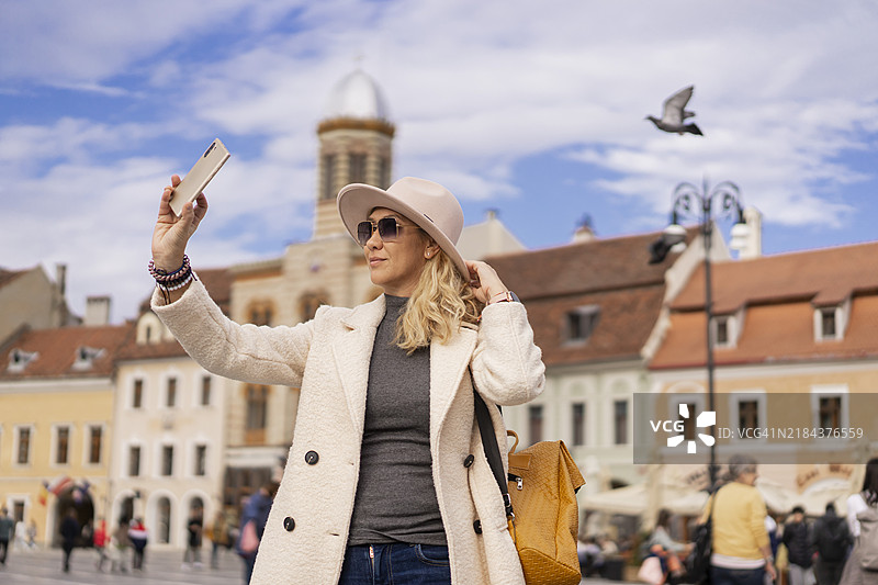 在罗马尼亚布拉索夫的欧洲广场上，一位戴着帽子的女性正在拍自拍，背景是复古建筑。图片素材