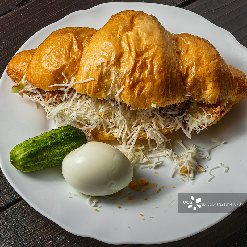 白盘上的金黄可颂三明治，夹有金枪鱼、鸡蛋、黄瓜和奶酪。图片素材