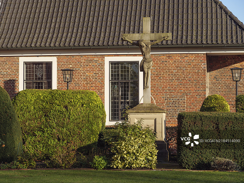 一座耶稣十字架矗立在砖墙前，周围是精心打理的花园和树篱，位于德国明斯特兰的布尔洛。图片素材