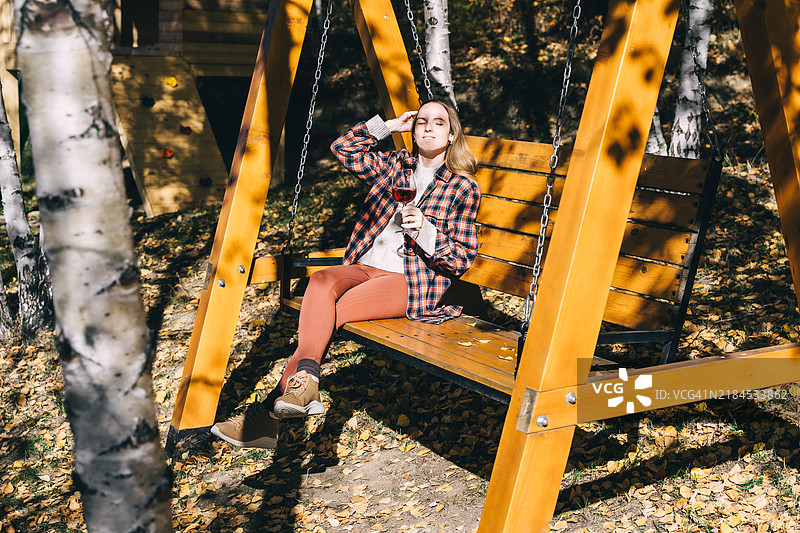 一位女士坐在木秋千上，手握红酒，享受秋天的美丽风景。图片素材