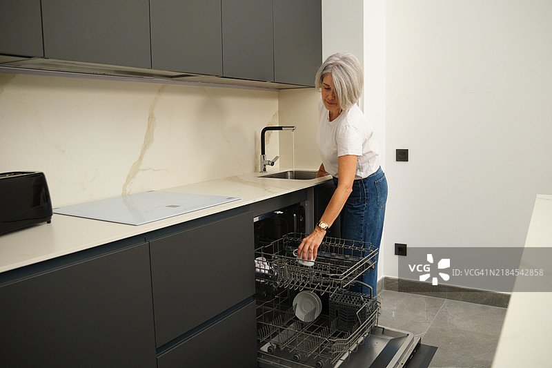 老年女性在现代厨房中将碗碟放入洗碗机图片素材