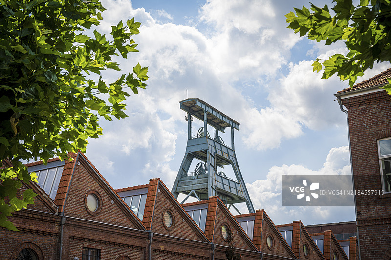 历史砖建筑上方的蜿蜒塔楼,四周环绕着绿色树木和蓝色多云的天空,赫尔滕的埃瓦尔德矿井蜿蜒塔。图片素材