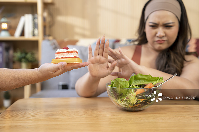 年轻的胖女人坐在厨房里，正在吃健康食品。她对甜食做出停止的手势，并且不看它。大码模特手里拿着一个番茄，右侧是健康食品。图片素材