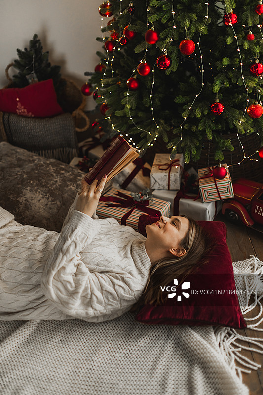 美丽的女人在家中圣诞树旁的垫子上放松地阅读书籍。图片素材