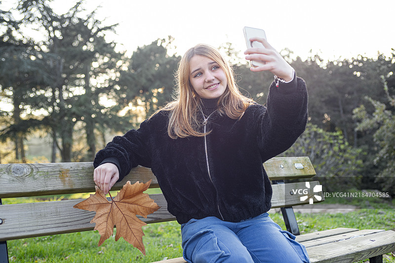 公园长椅上坐着的少女用智能手机自拍。图片素材