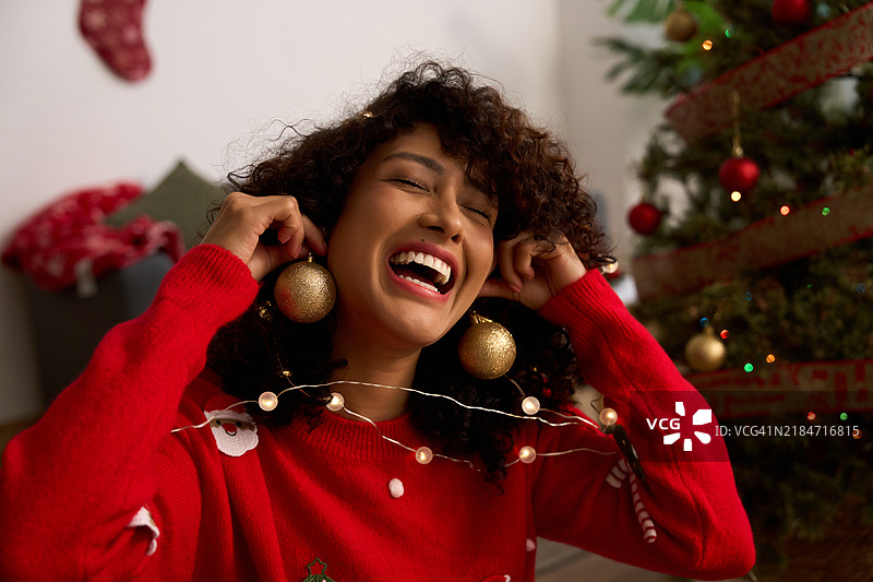 年轻女性佩戴圣诞饰品耳环，笑着站在圣诞树旁图片素材