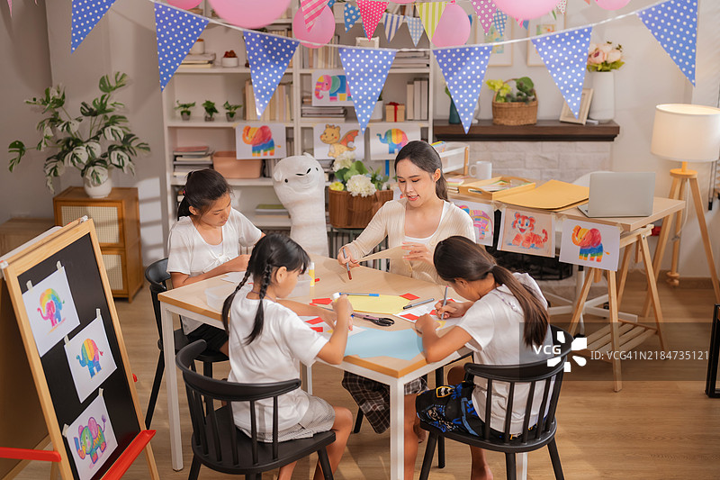 亚洲美丽女性教师正在向一群专注的学生讲解如何进行剪纸和拼贴艺术。美丽的教师在日间托儿所与孩子们一起享受学习的乐趣。图片素材