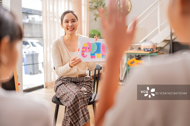 美丽的亚洲女老师在幼儿园级别为学生们解释和教授作业的意义，向全班展示作业纸拼贴，愉快地享受在家庭托儿所的课堂。图片素材