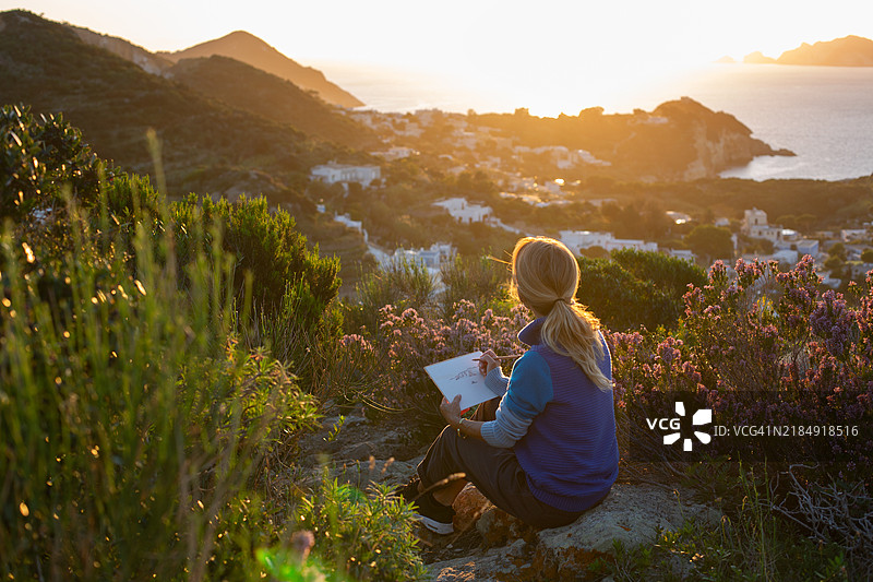 日落时分，女人坐在山顶上画画。图片素材