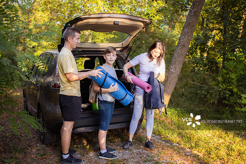 快乐的家庭为冒险做准备。母亲、父亲和儿子正在从汽车后备箱中拿东西，准备去大自然中徒步旅行或野餐。图片素材