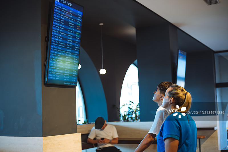 一对情侣在机场焦急地等待，目光紧盯着出发信息牌，满怀期待地等待即将开始的旅程。这张图片捕捉了旅行的兴奋与不确定性，非常适合与机场、假期相关的主题。图片素材