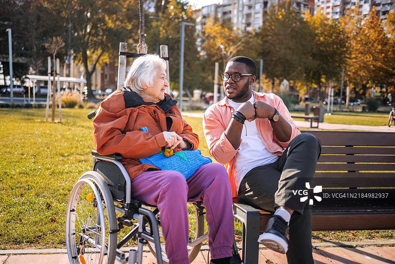 年轻志愿者在公园长椅上与坐在轮椅上的老人交谈，关心他们的户外活动。图片素材