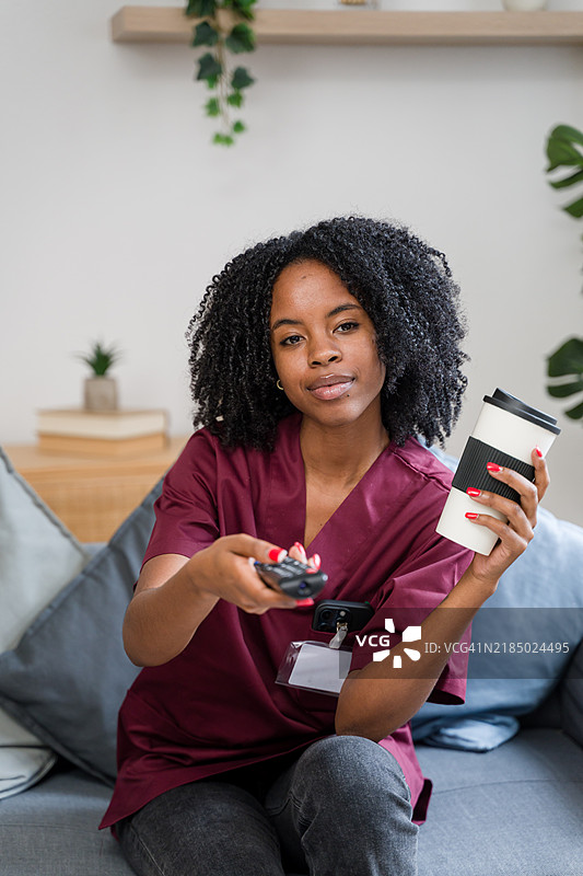 护士下班后在沙发上放松，手里拿着咖啡和遥控器。图片素材