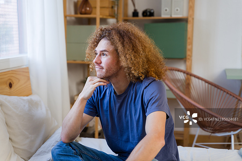 年轻男子坐在卧室的床上，思考着，窗边静静地望着外面。图片素材