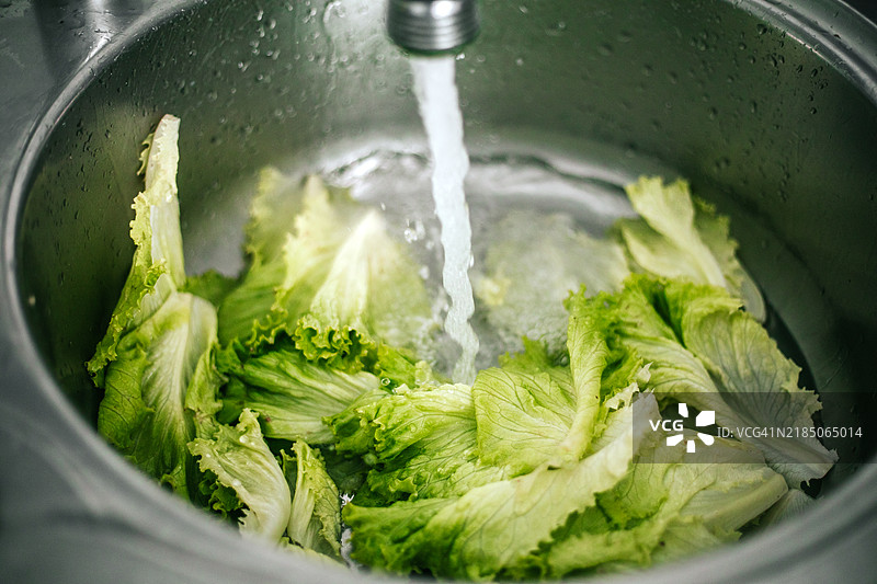 一束新鲜的绿叶生菜正在厨房水槽中清洗，水流淌过鲜艳的绿色叶子。生菜被轻轻冲洗以去除任何污垢。图片素材