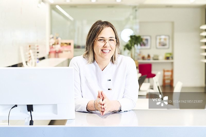 穿着白大褂的女性验光师微笑着看向镜头，靠在办公室的电脑旁，正面视图图片素材