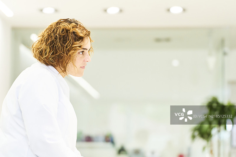 身穿实验室外套的女性验光师在眼镜店侧身站立，目光投向一旁。图片素材