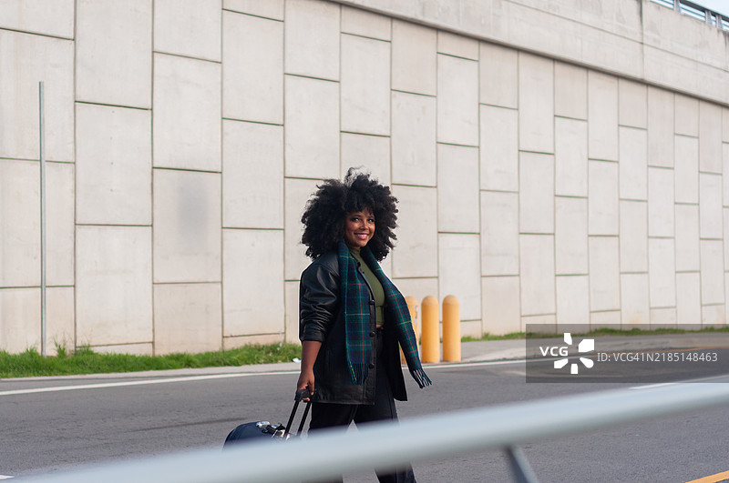 一位非洲裔女性开心地走在城市中，拖着行李箱，准备探索和迎接她的旅程。图片素材