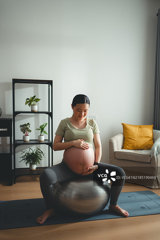 亚洲孕妇坐在健身球上，轻松地在家中抚摸着自己的肚子。图片素材