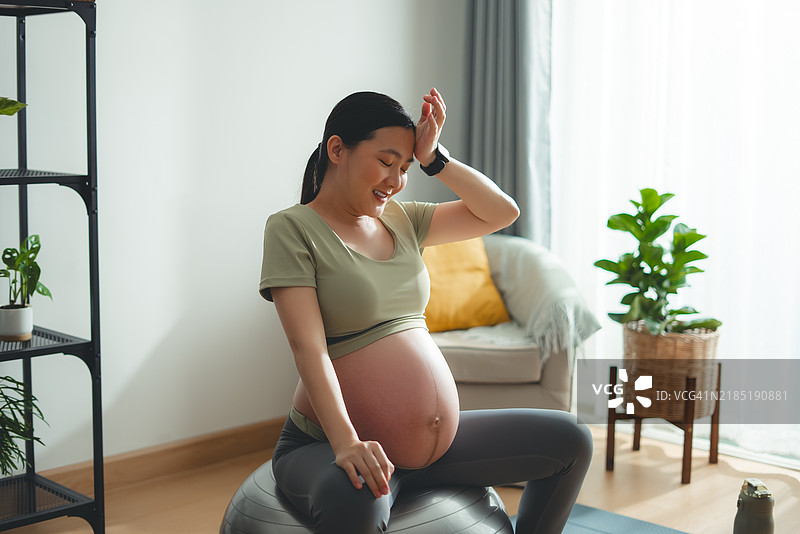 亚洲孕妇在家中客厅坐在健身球上，擦去汗水，放松心情，开心地微笑，刚刚锻炼完。图片素材