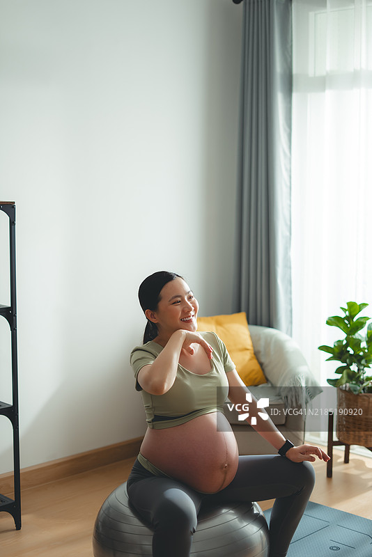 亚洲孕妇在家中客厅坐在健身球上，擦去汗水，放松心情，开心地微笑，刚刚锻炼完。图片素材