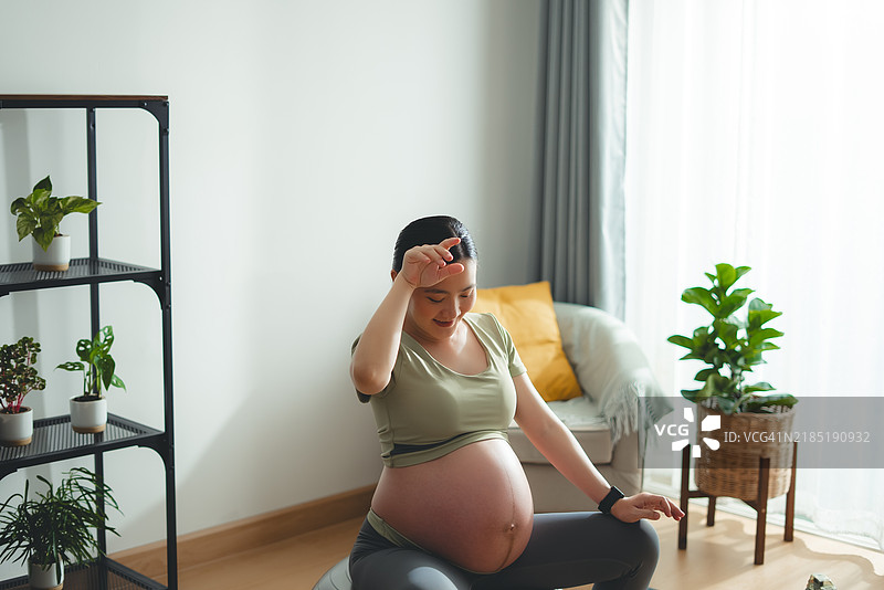 亚洲孕妇在家中客厅坐在健身球上，擦去汗水，放松心情，开心地微笑，刚刚锻炼完。图片素材