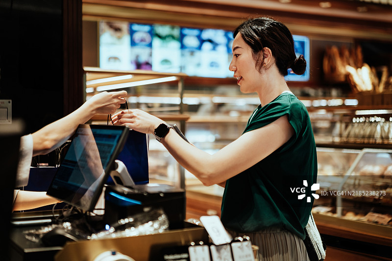 年轻的亚洲女性顾客在商店的收银台从店主那里拿到纸袋。顾客购物体验。当地商家的温暖友好的服务。图片素材