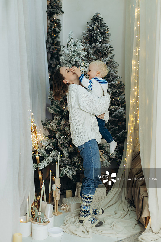 年轻女性抱着一个顽皮的男孩，背景是装饰华丽的新年树，置身于节日的房间里。图片素材