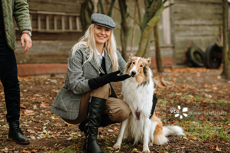 时尚的秋日。一位穿着粗花呢夹克和帽子的女性在乡村环境中与她的粗毛牧羊犬享受美好时光。这个画面唤起了优雅、陪伴和秋天的美丽。非常适合生活方式、时尚和宠物相关的项目。图片素材