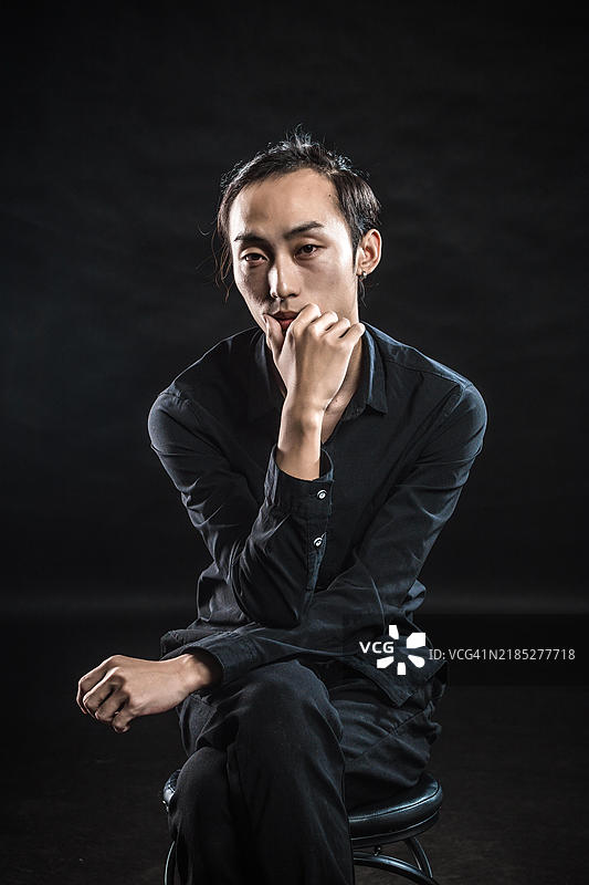 一位年轻的亚洲男子穿着黑色衬衫和裤子，坐在椅子上，手托着下巴。图片素材