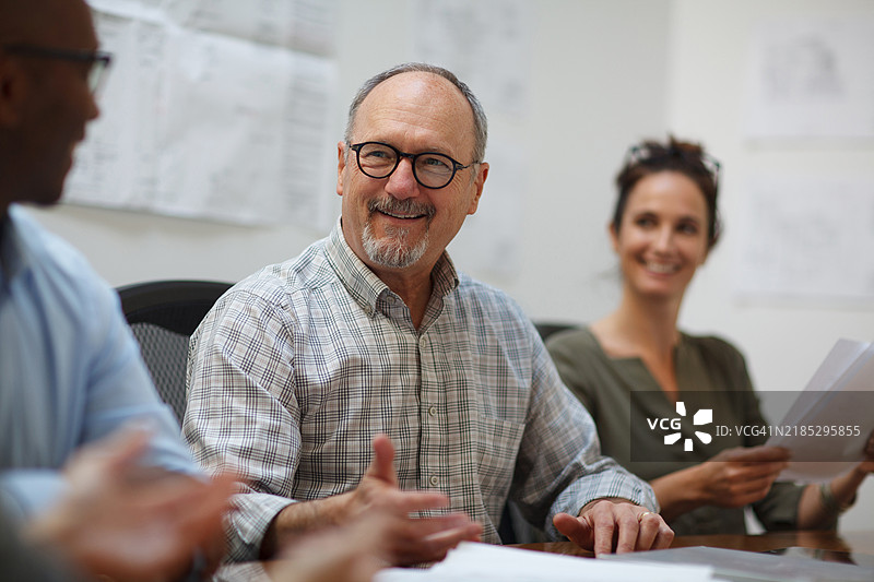 一位年长男性在设计办公室的会议中与同事们微笑交谈，坐在桌子旁。图片素材