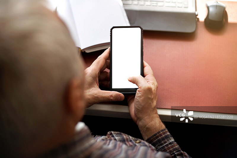 老年人坐在家里的桌子旁，肩膀后方视角使用智能手机。图片素材