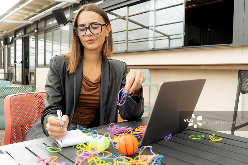 年轻女性穿着黑色皮夹克，使用电脑，做笔记，并利用橡皮筋、玩具和辅助设备来提高专注力。图片素材